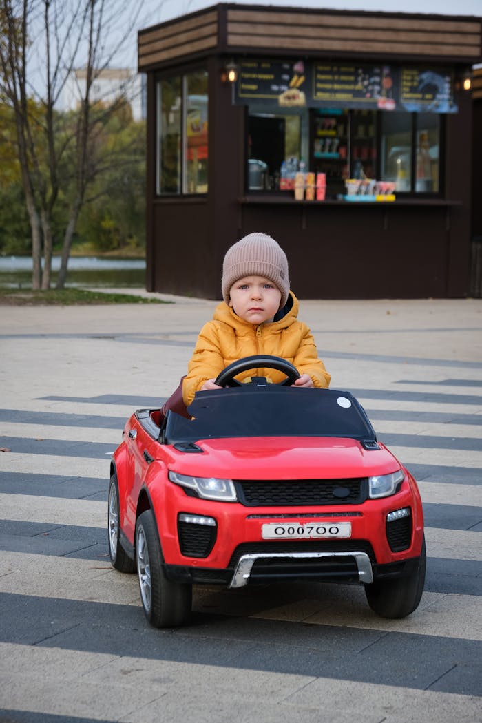 Young child in a red toy car at a city park, dressed in winter clothing on a sidewalk.